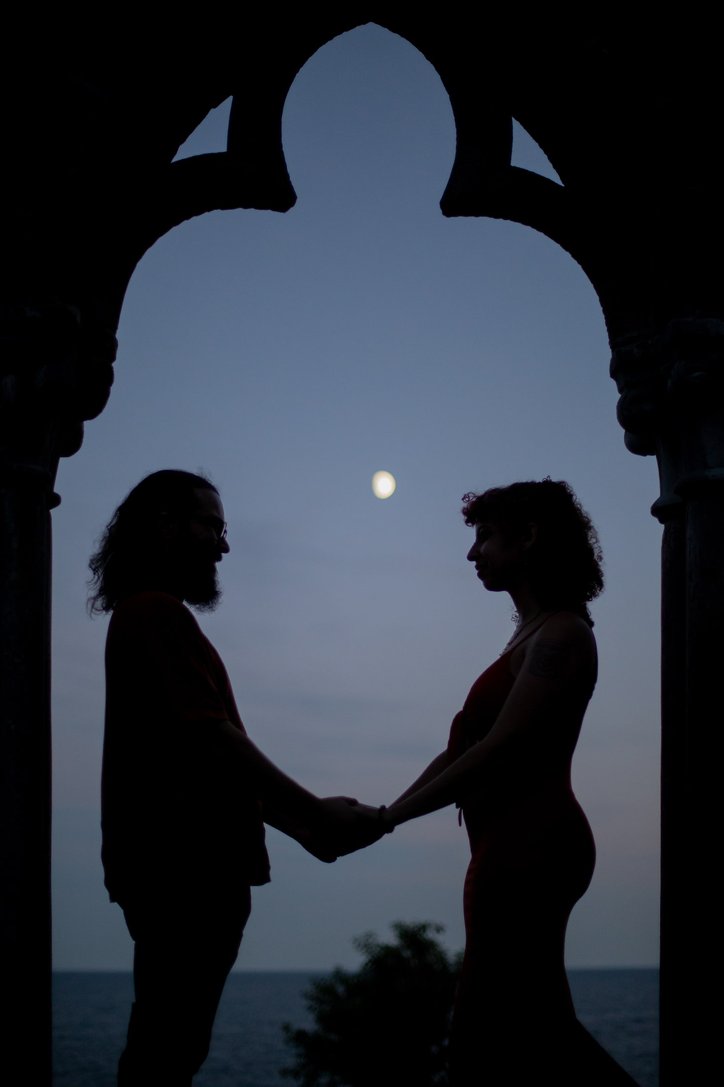 A man and woman hold hands as they stand above the ocean, framed by a stone arch, between them the moon, photographed at the blue hour during their wedding proposal photoshoot at Hammond Castle Museum by Boston wedding photographers Spagnolo Photography.