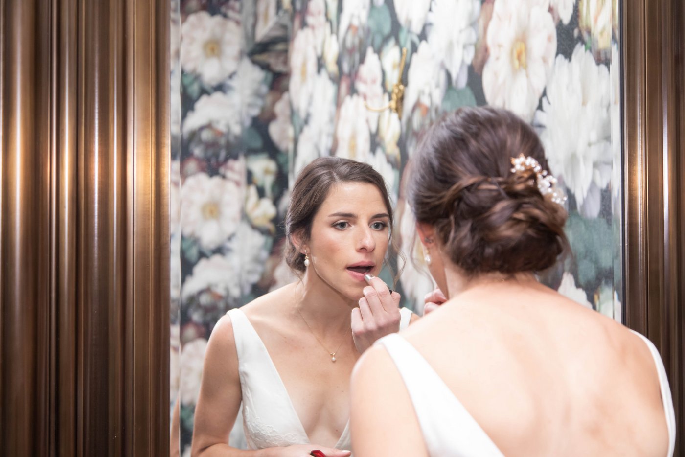 Bride applying lipstick against floral wallpaper before her fall wedding in Brookline