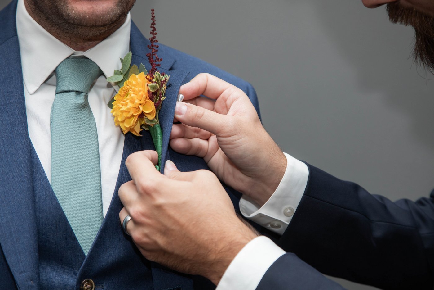 A close up of a groomsman's hands fastening a boutonniere on the groom's lapel