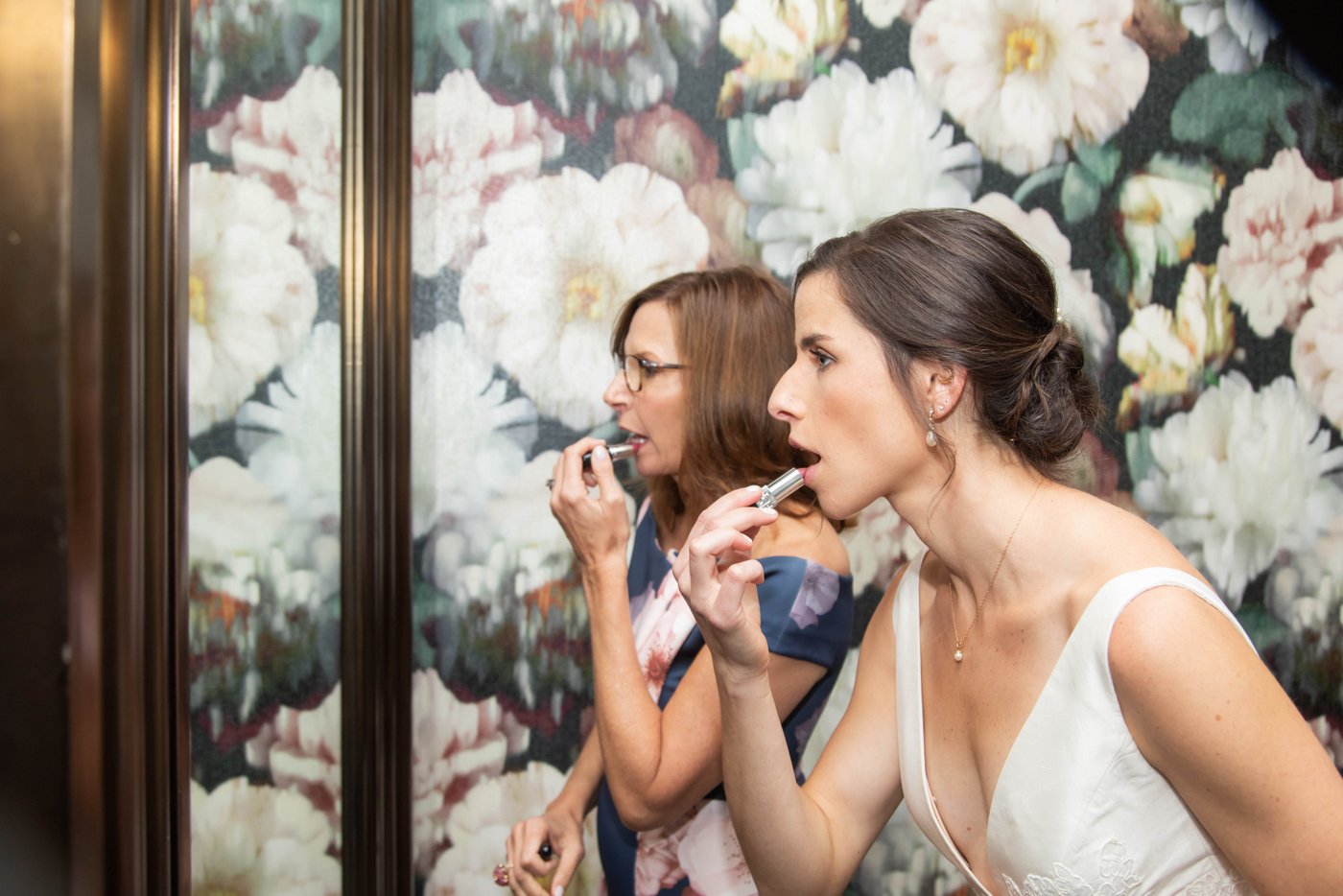 Bride and her mother applying lipstick against floral wallpaper before her fall wedding in Brookline.