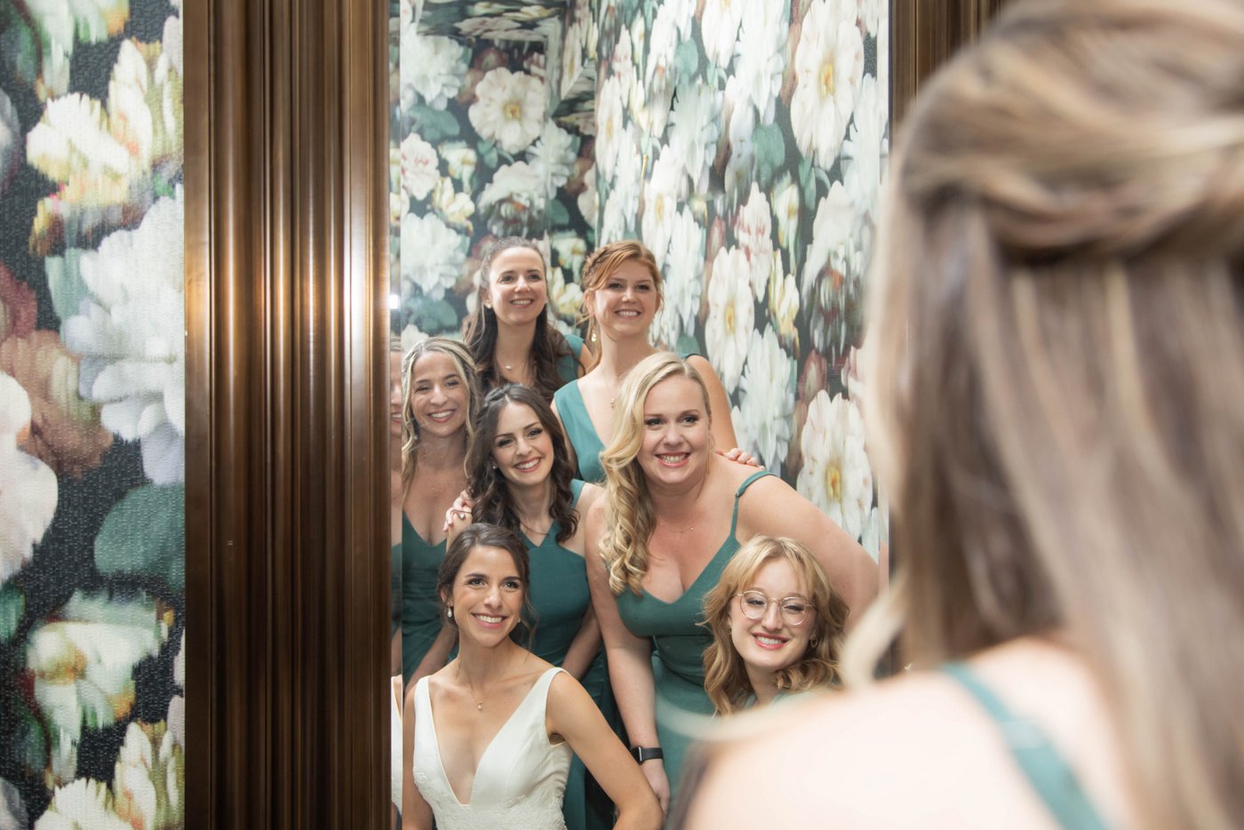 Bride and bridesmaid looking in a mirror in a room with floral wallpaper before her fall wedding in Brookline