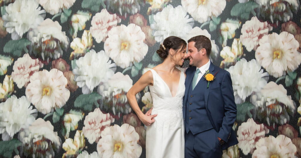 Bride and groom grinning at each other while standing against a floral wallpaper before their fall wedding in Brookline, MA, photographed in a natural candid style by creative Boston wedding photographers Spagnolo Photography