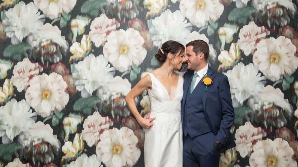 Bride and groom grinning at each other while standing against a floral wallpaper before their fall Brookline Jewish wedding, photographed in a natural candid style by creative Boston wedding photographers Spagnolo Photography