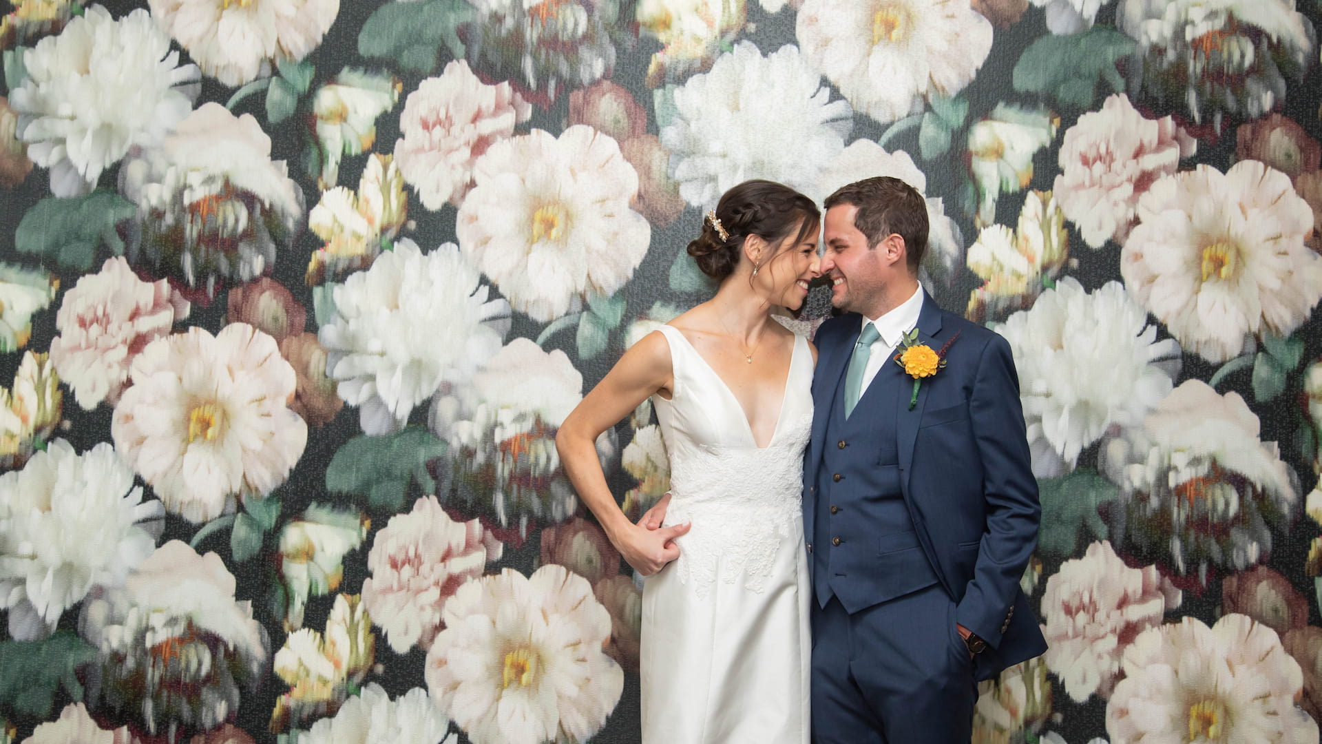 Bride and groom grinning at each other while standing against a floral wallpaper before their fall Brookline Jewish wedding, photographed in a natural candid style by creative Boston wedding photographers Spagnolo Photography