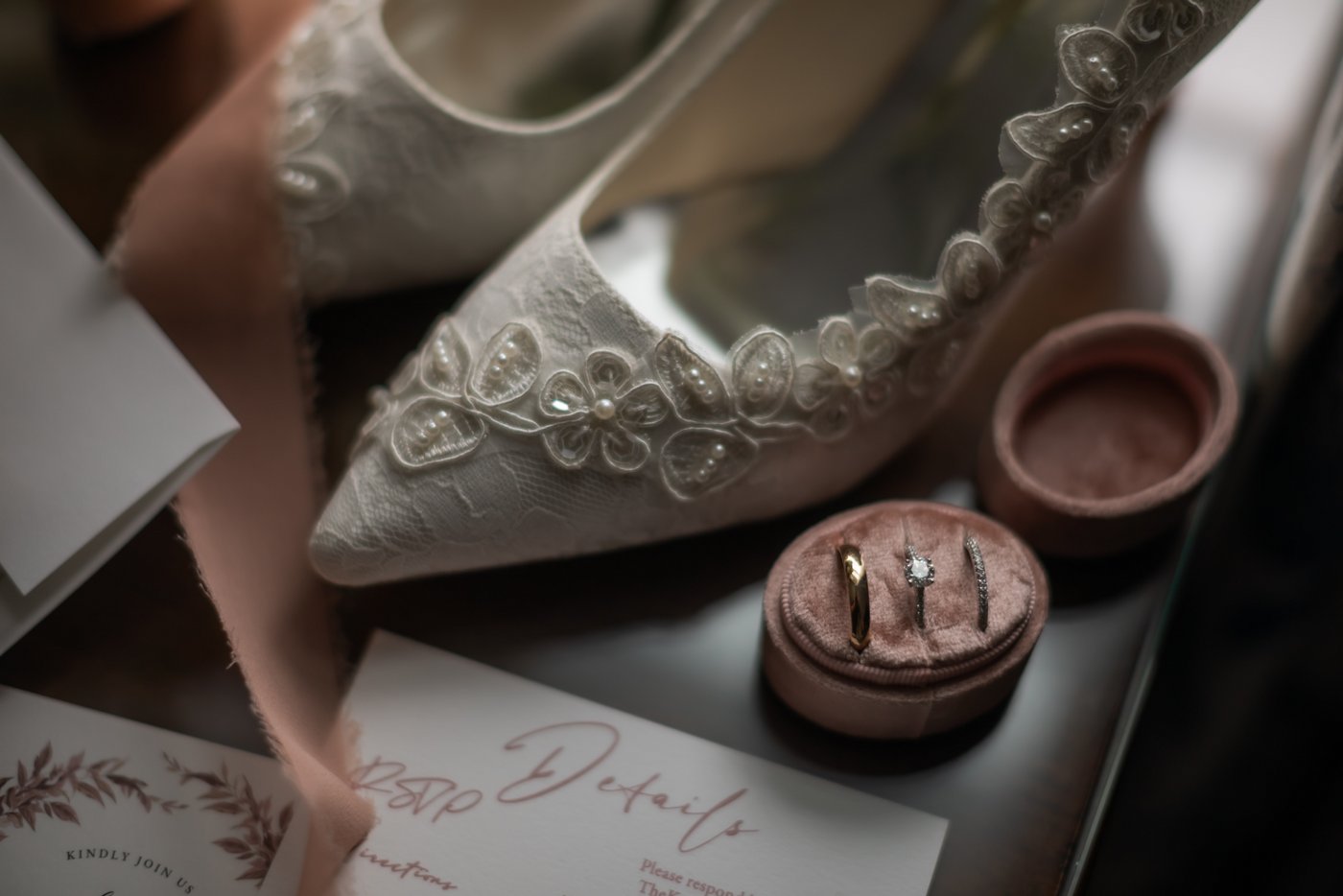 The wedding bands in a pink velvet ring holder, next to the bride's lacy wedding shoes with pearl details, photographed at the Eliot Hotel in Boston