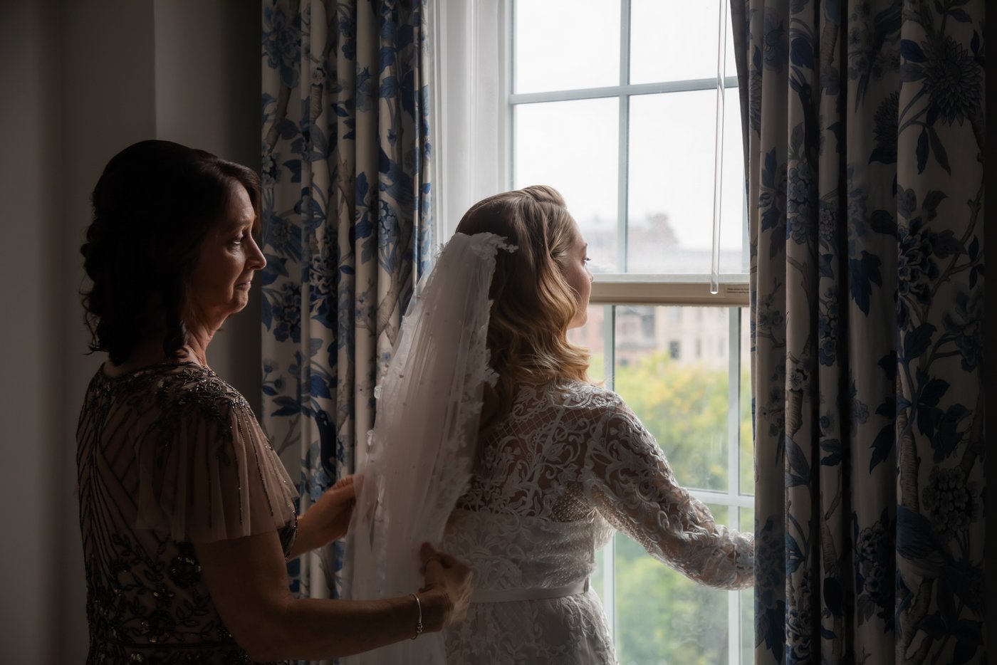 A moody photo of a bride looking out of the window of her hotel suite as her mother adjusts her veil, photographed at the Eliot Hotel in Boston by Boston wedding photographers Spagnolo Photography