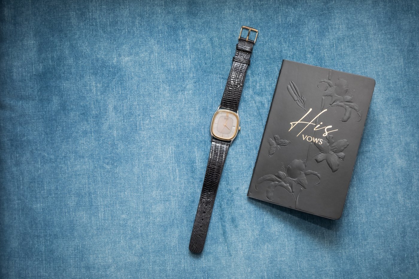 The groom's vow book and his gold-rimmed watch with black leather straps, against a blue canvas