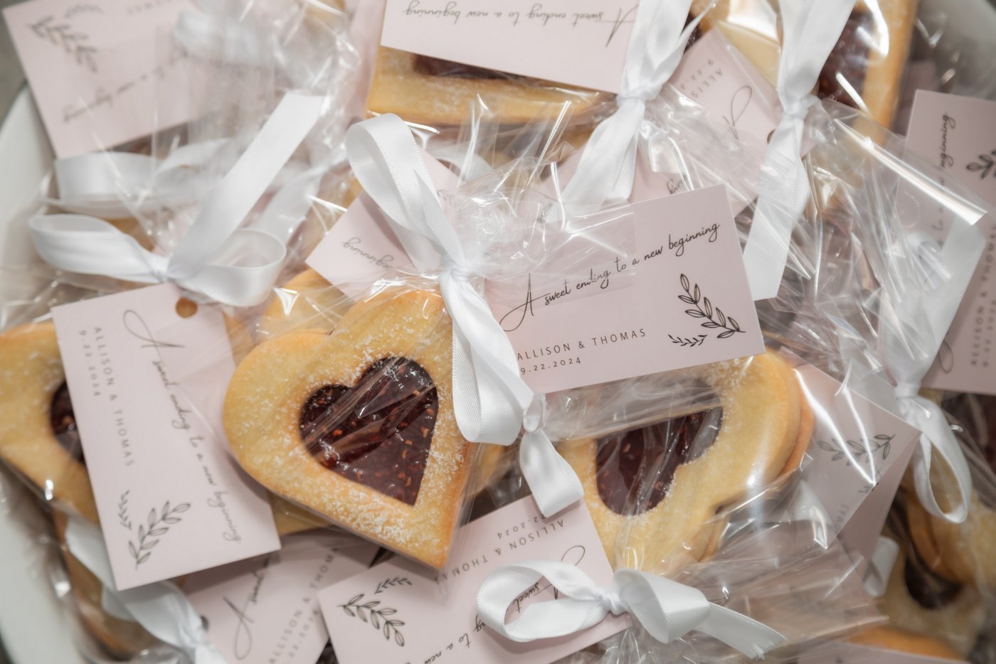 Heart shaped jam-filled Linzer cookies individually wrapped in clear bags and tied with white ribbons with a tag that reads "A sweet ending to a new beginning," to be gifted to guests as wedding favors