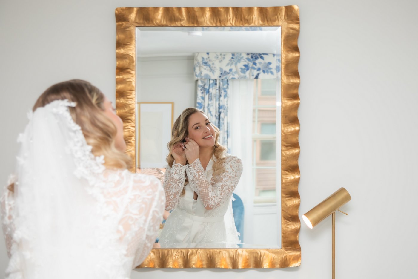 The bride adjusting her earrings while looking at the camera through the mirror on the wall, photographed in the bridal suite of the Eliot Hotel in Boston