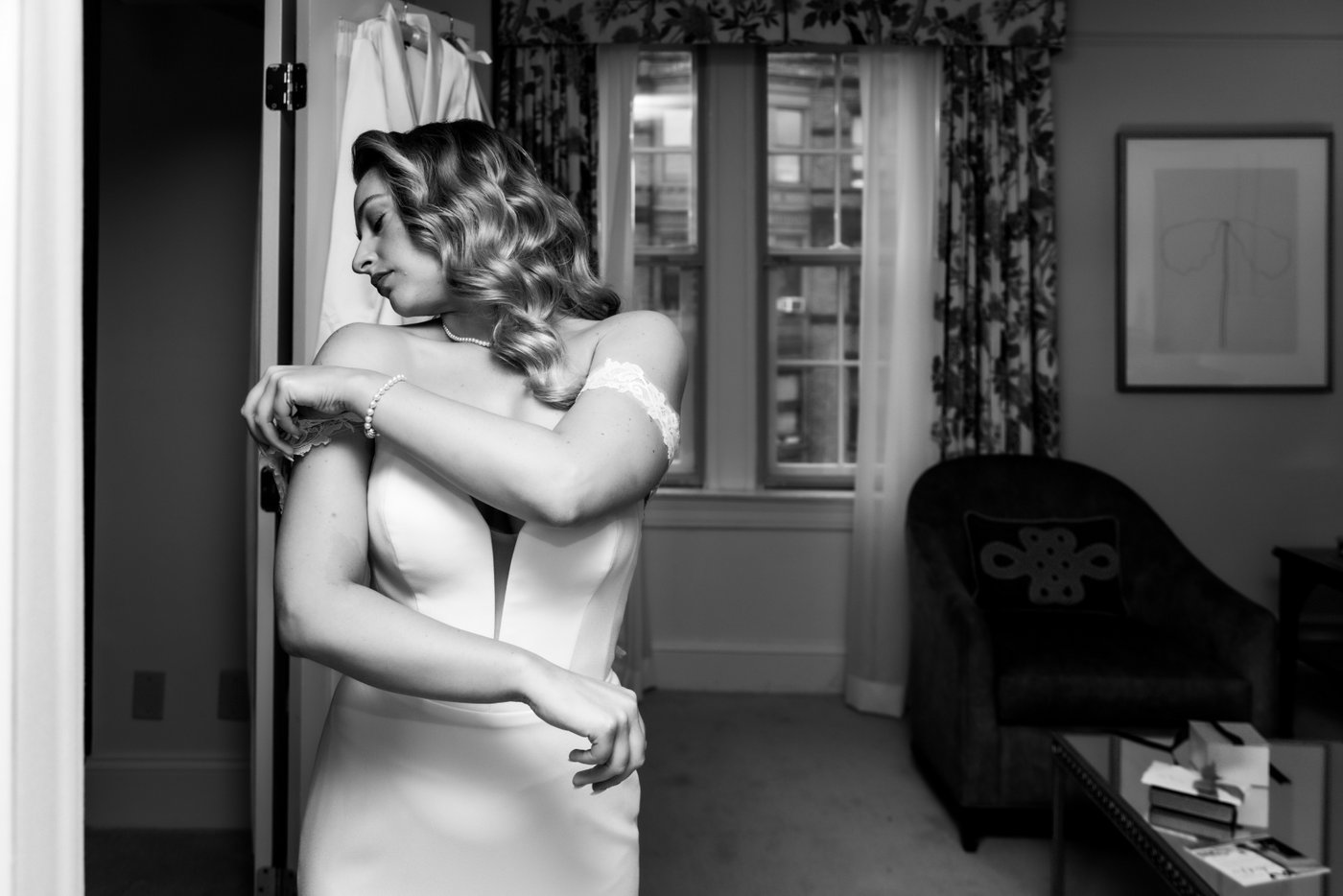 An elegant black and white portrait of a bride adjusting the straps of her wedding dress in her bridal suite at the Eliot Hotel in Boston, where she got ready for her Terra at Eataly wedding, photographed by Boston wedding photographers Spagnolo Photography