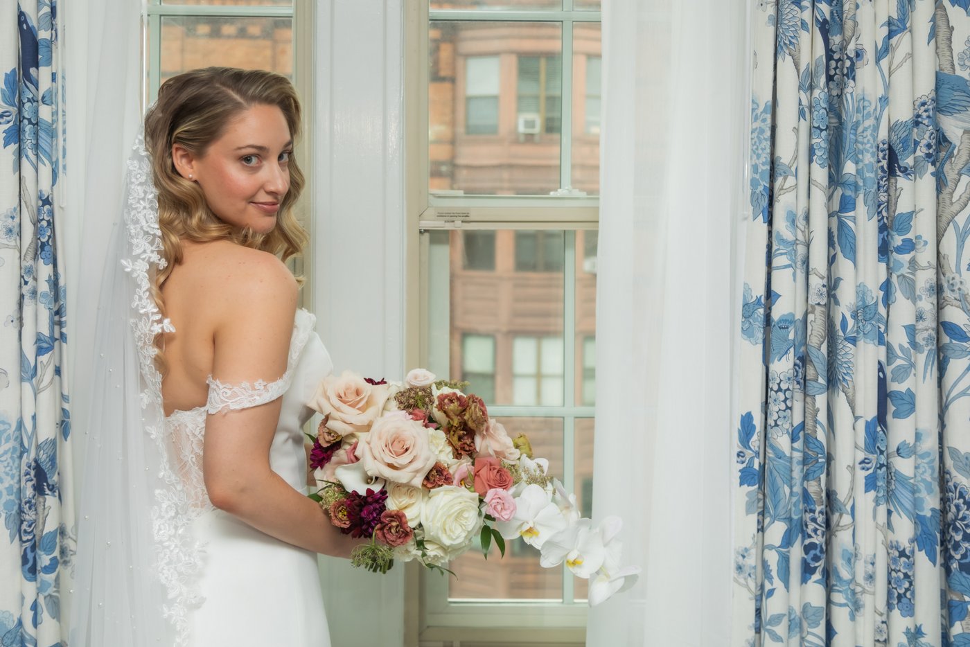 A portrait of the bride holding her large bouquet of flowers and looking back at the camera, as she stands by the window of her bridal suite at the Eliot Hotel in Boston