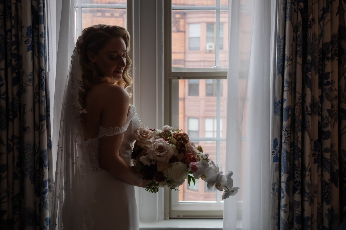 A moody portrait of a bride holding a large bouquet of flowers standing by the window in her bridal suite at the Eliot Hotel in Boston on her wedding day.