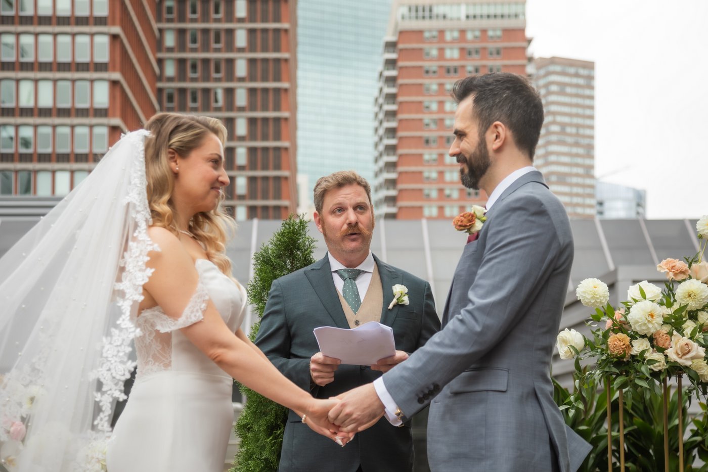 A bride and a groom hold hands and smile as the officiant speaks during their wedding ceremony on the rooftop of Terra at Eataly in Boston where they held their micro wedding, photographed by Boston wedding photographers Spagnolo Photography