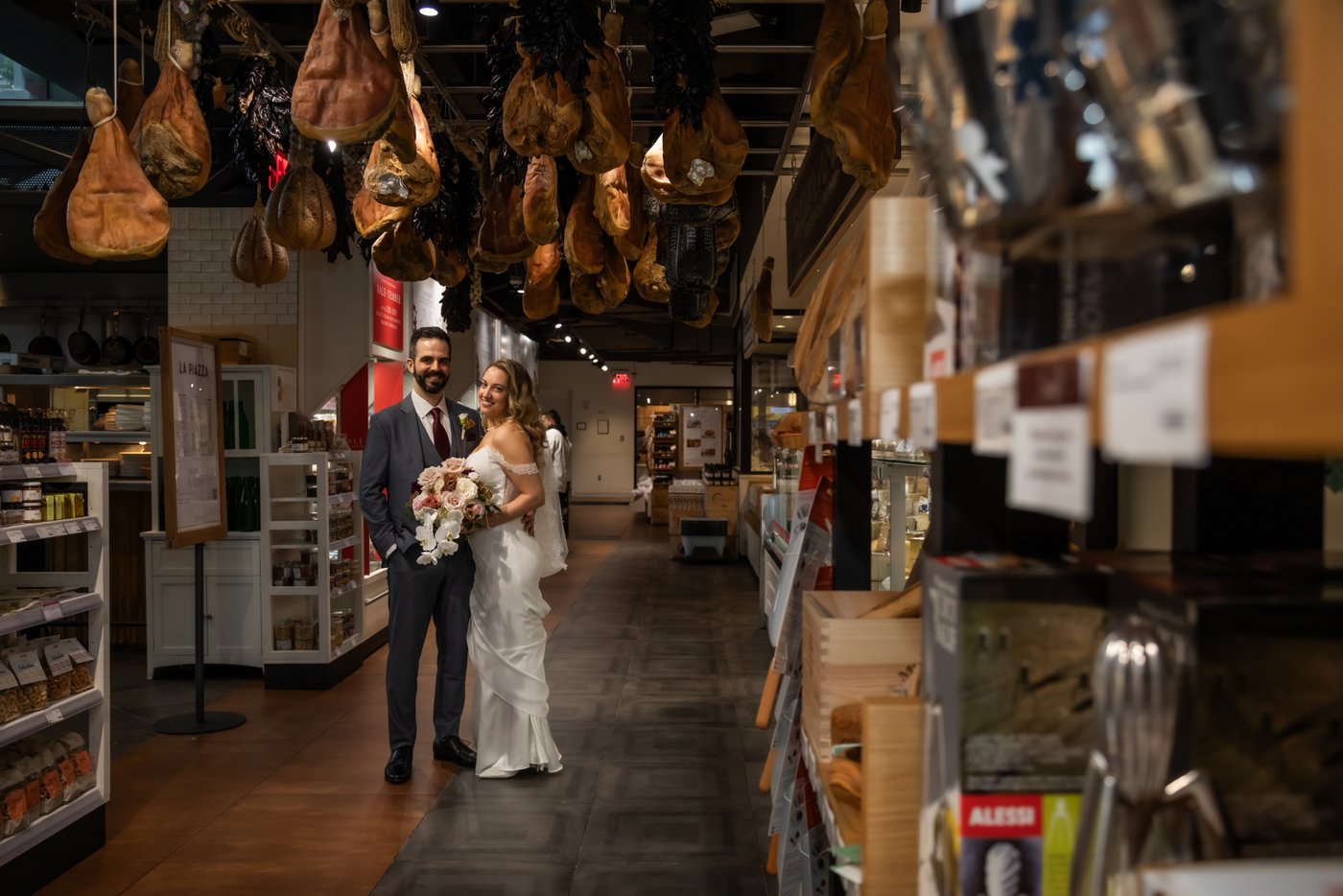 A bride and groom stand under cured meats hanging from the ceiling at Eataly in Boston, photographed by Spagnolo Photography for their micro-wedding photography package.