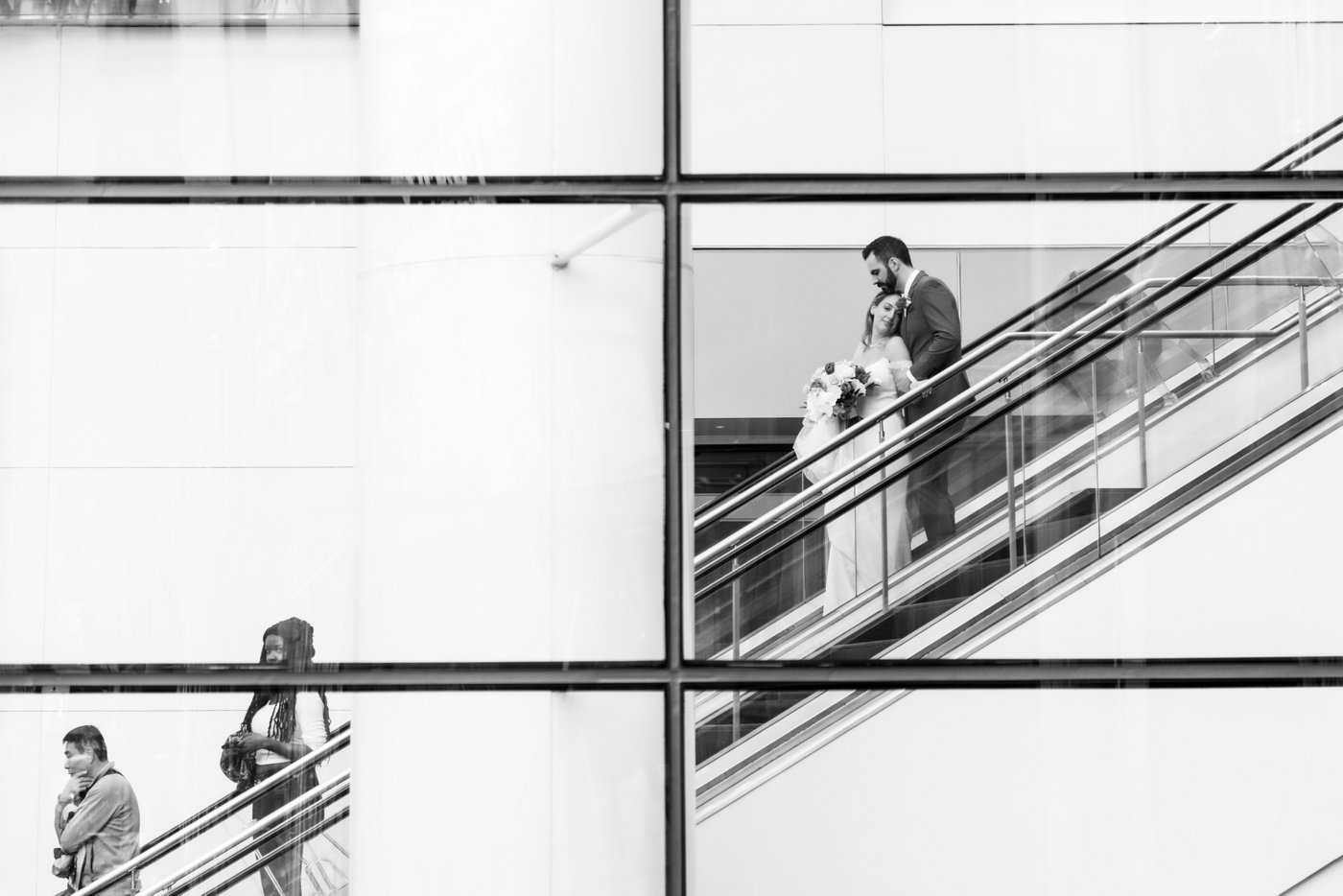 A black and white photo of a bride and a groom riding down the escalators at Boston's Prudential Center, during their wedding at Terra at Eataly