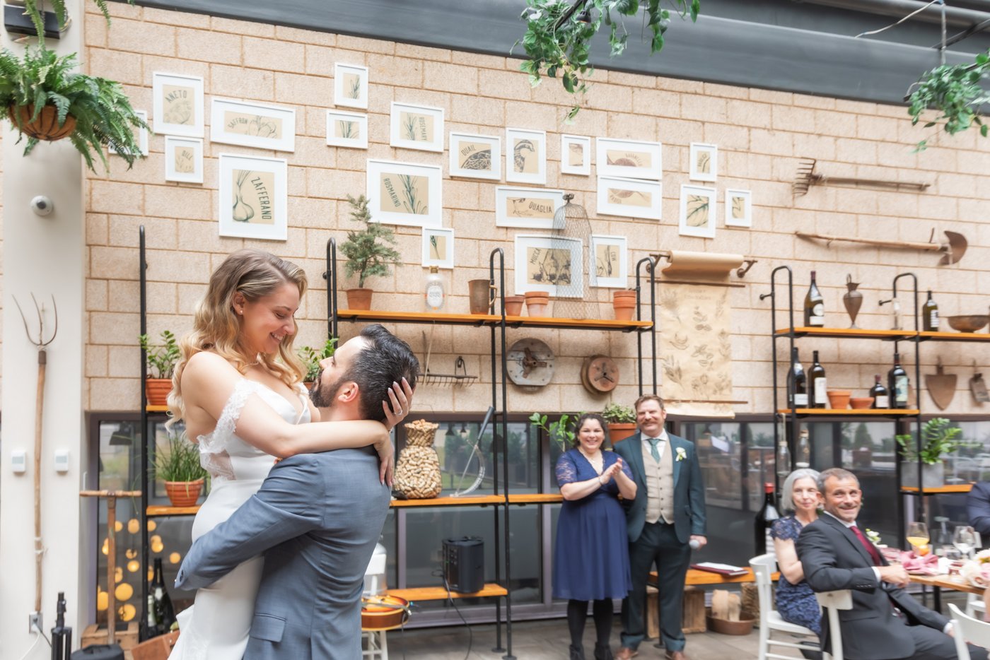 The groom picks up the bride during their first dance while guests watch near a wall decorated with potted plants, paintings, and gardening tools at a Terra at Eataly wedding in Boston