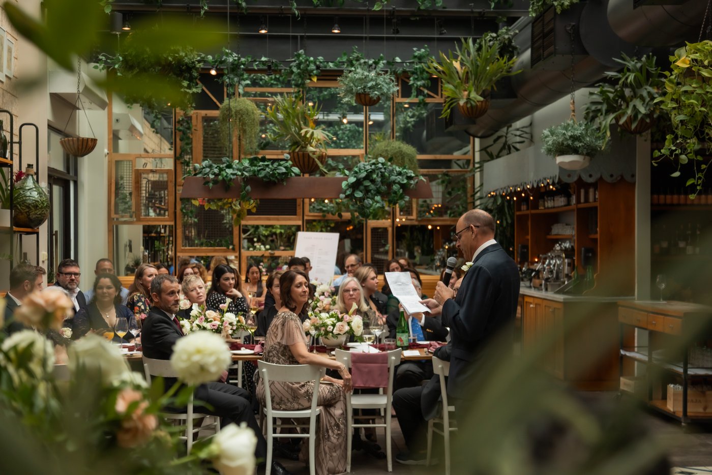 Micro-wedding guests at Terra at Eataly listen to a speech surrounded by lush green hanging plants.
