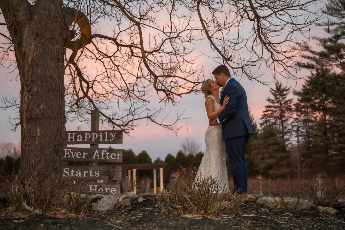 A bride and a groom kissing at sunset with a pink sky behind then at Birch Wood Vineyards on their wedding day, beside them a sign reads, "Happily Ever After Starts Here," photographed by Spagnolo Photography in 2025 in Derry, New Hampshire.