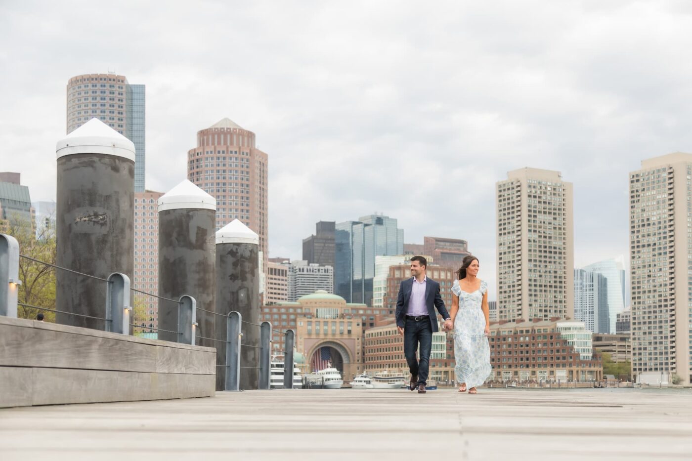 A couple walks on the boardwalk of the Boston Harbor Walk during their engagement photography session with Spagnolo Photography