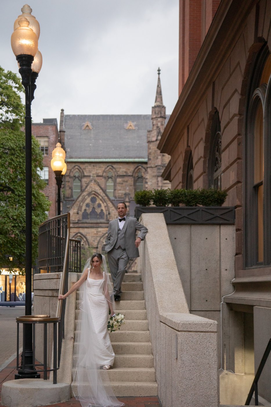 A bride and a groom stand on the steps of an old Boston building, next to city lamps with amber glows, photographed at sunset during a Boston wedding by Boston wedding by Spagnolo Photography.