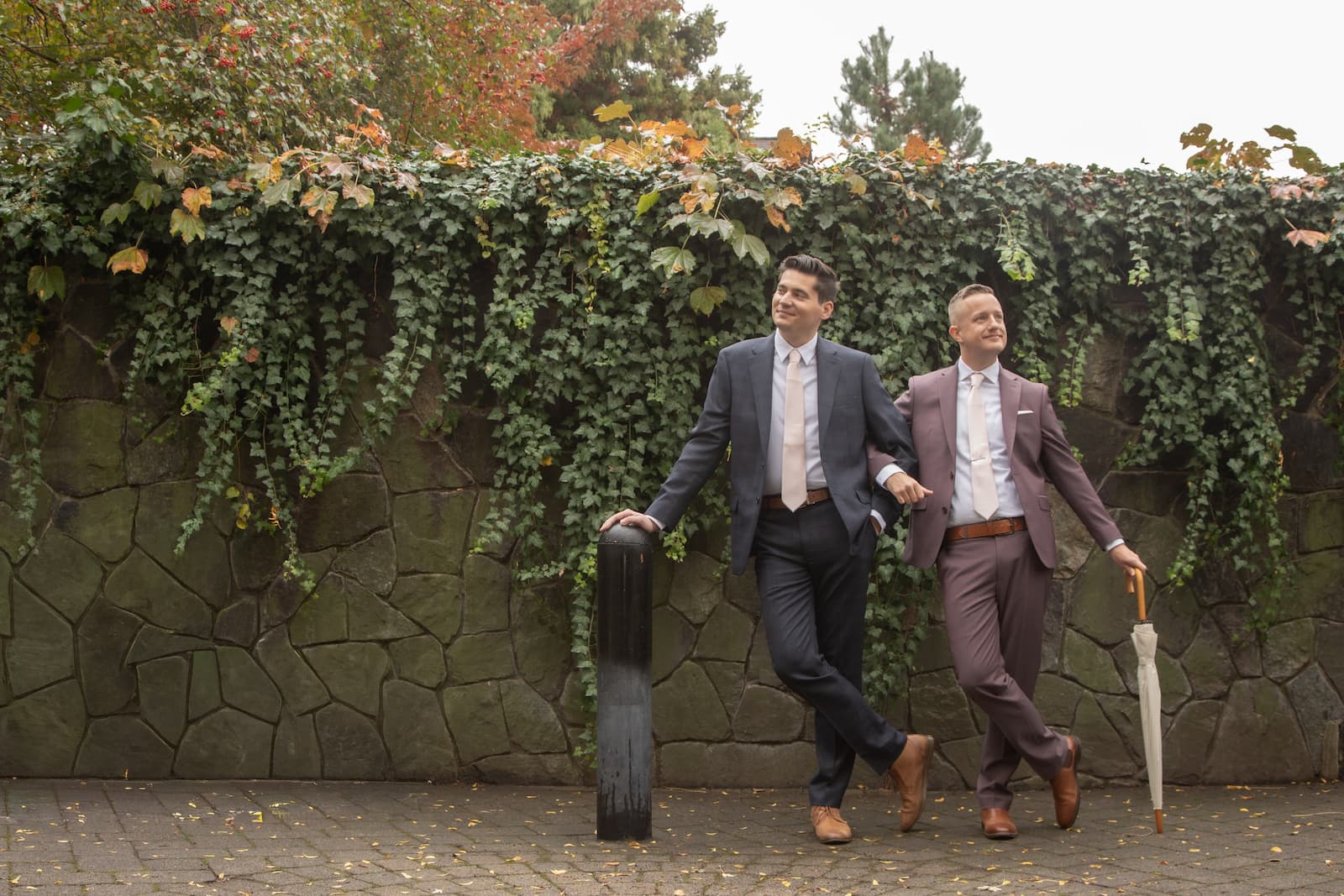 Two grooms posing while linking arms in front of a stone wall covered in ivy, photographed at Cambridge City Hall just before their wedding ceremony.