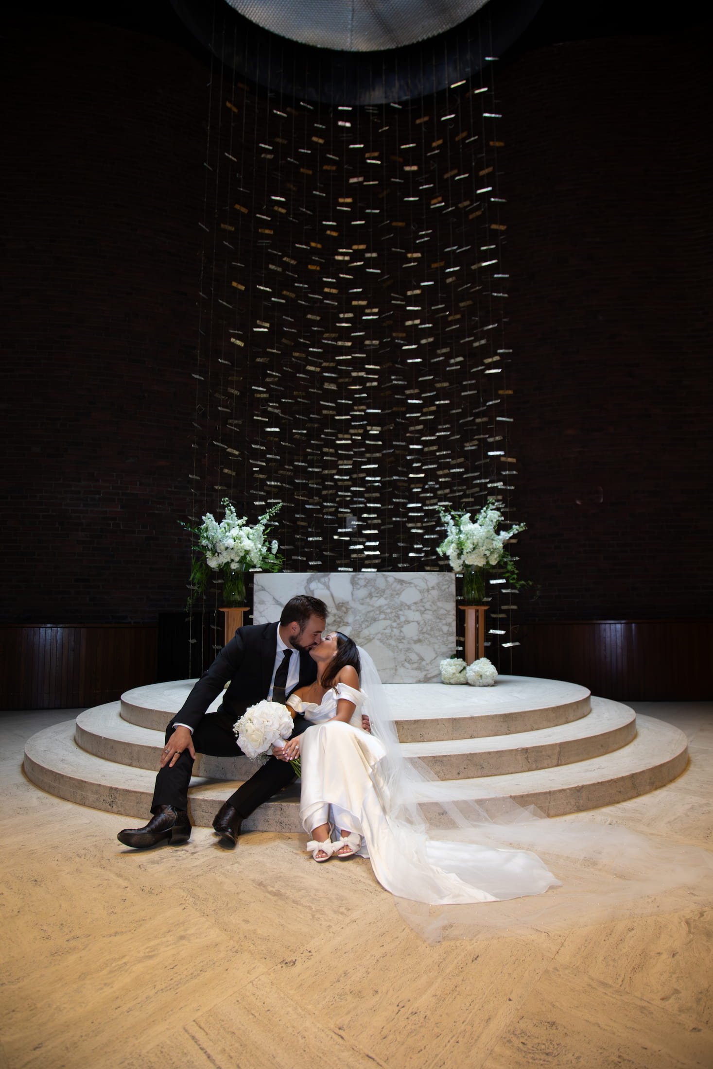 A bride and groom kissing while sitting on the stairs of the altar at the MIT Chapel after their wedding ceremony in the summer 2025