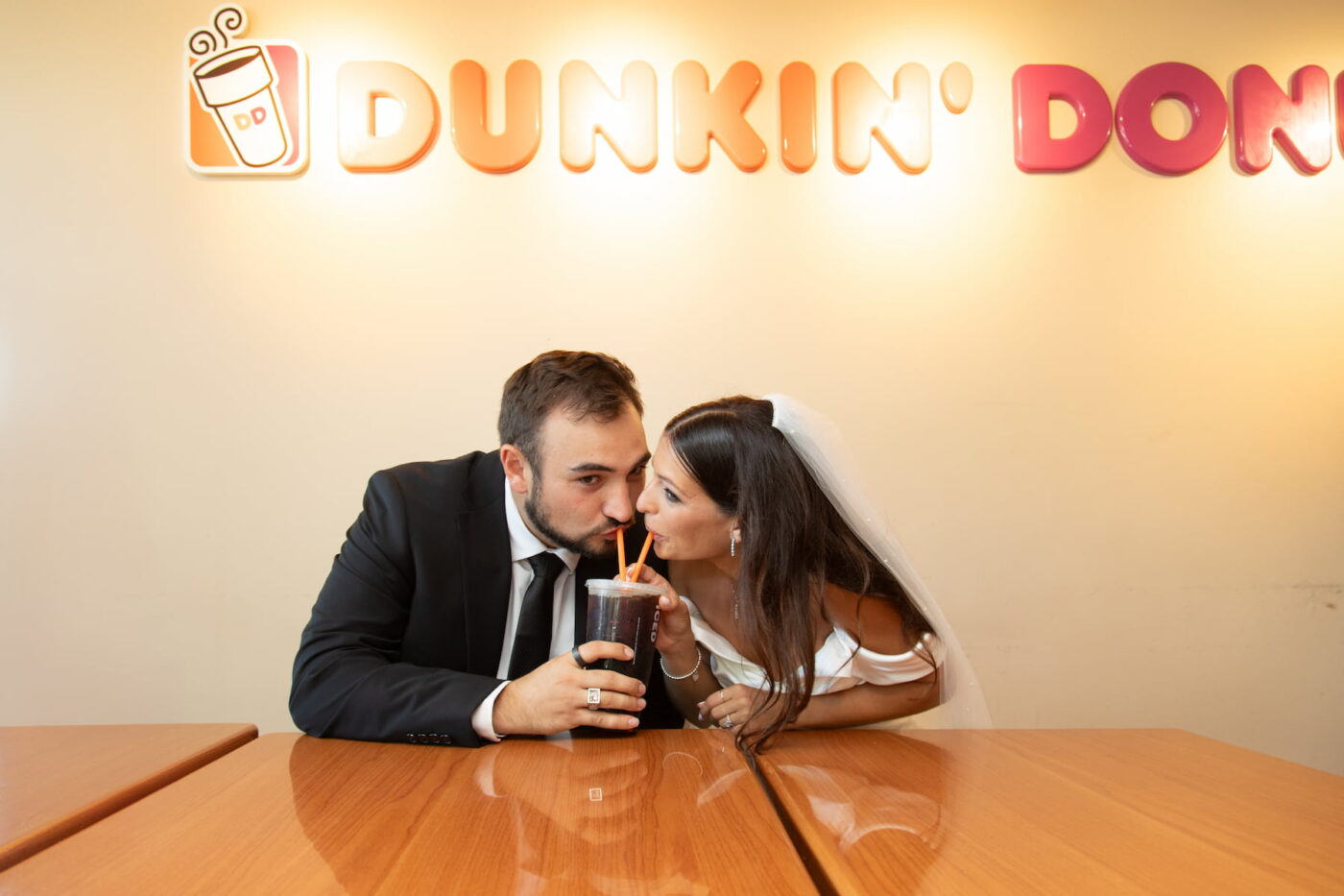 A bride and a groom share an iced coffee with two straws at a Dunkin' Donuts during their MIT wedding, photographed by Boston wedding photographers Spagnolo Photography