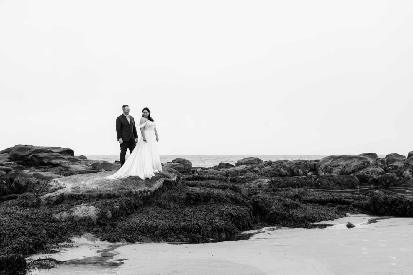 A black and white photo of a bride and a groom standing on rocks on Wingaersheek Beach during their beach wedding portraits