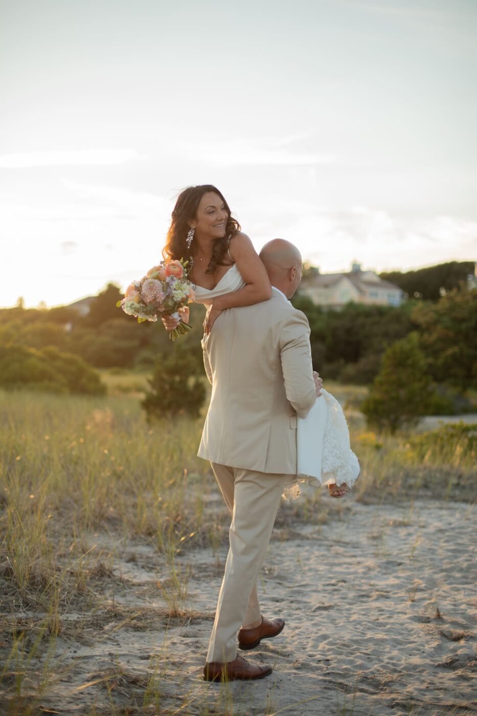 A groom carries his bride over his shoulder as he walks through the dunes at sunset during their Wychmere Beach Club wedding in Harwich Port, MA