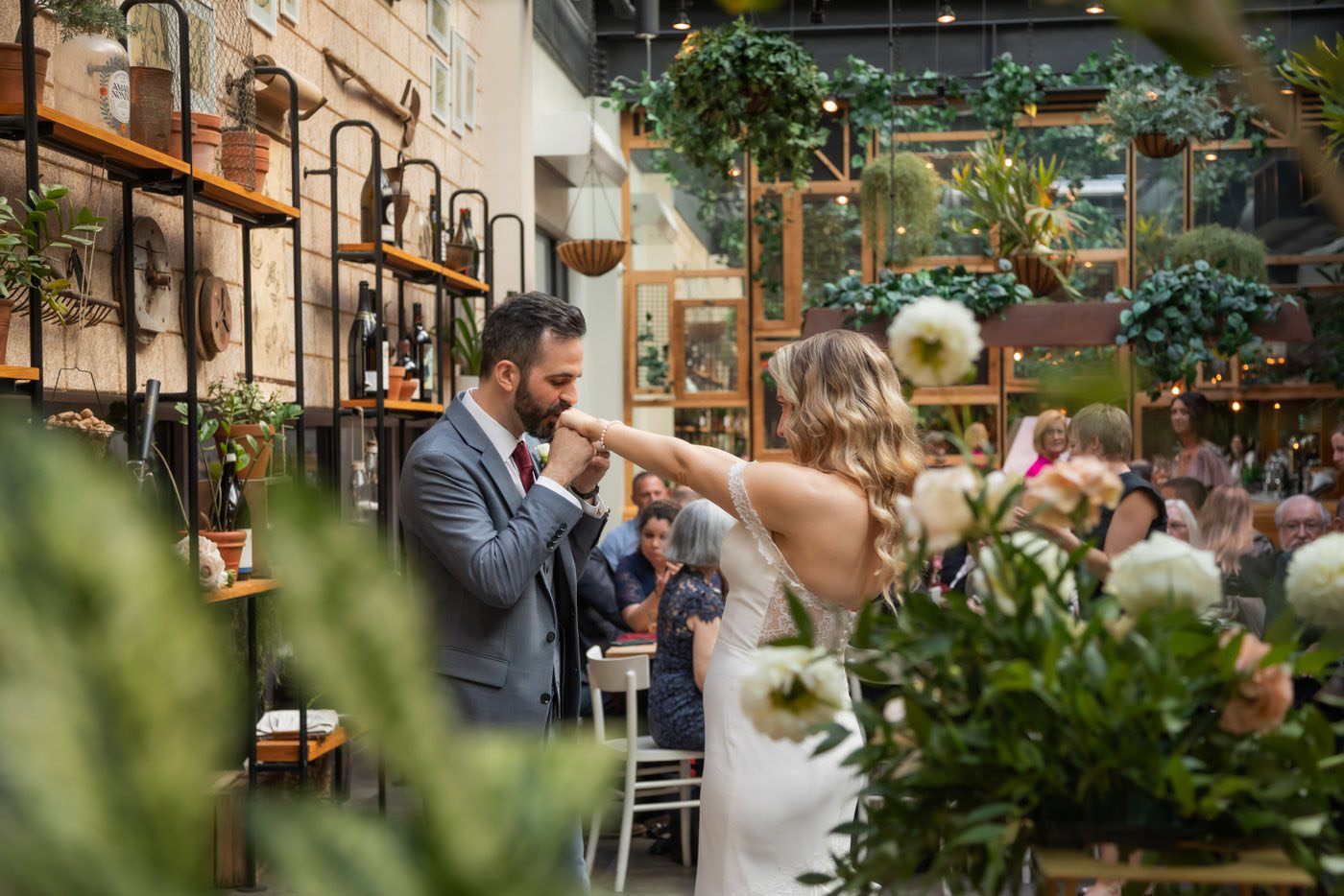 The groom kissing the bride's hand as they are surrounded by lush green plants and loved ones at a Terra Boston wedding