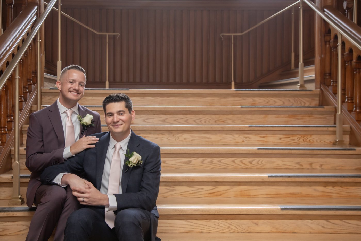 Two grooms smile at the camera as they sit on the interior stairs of Cambridge City Hall on their wedding day