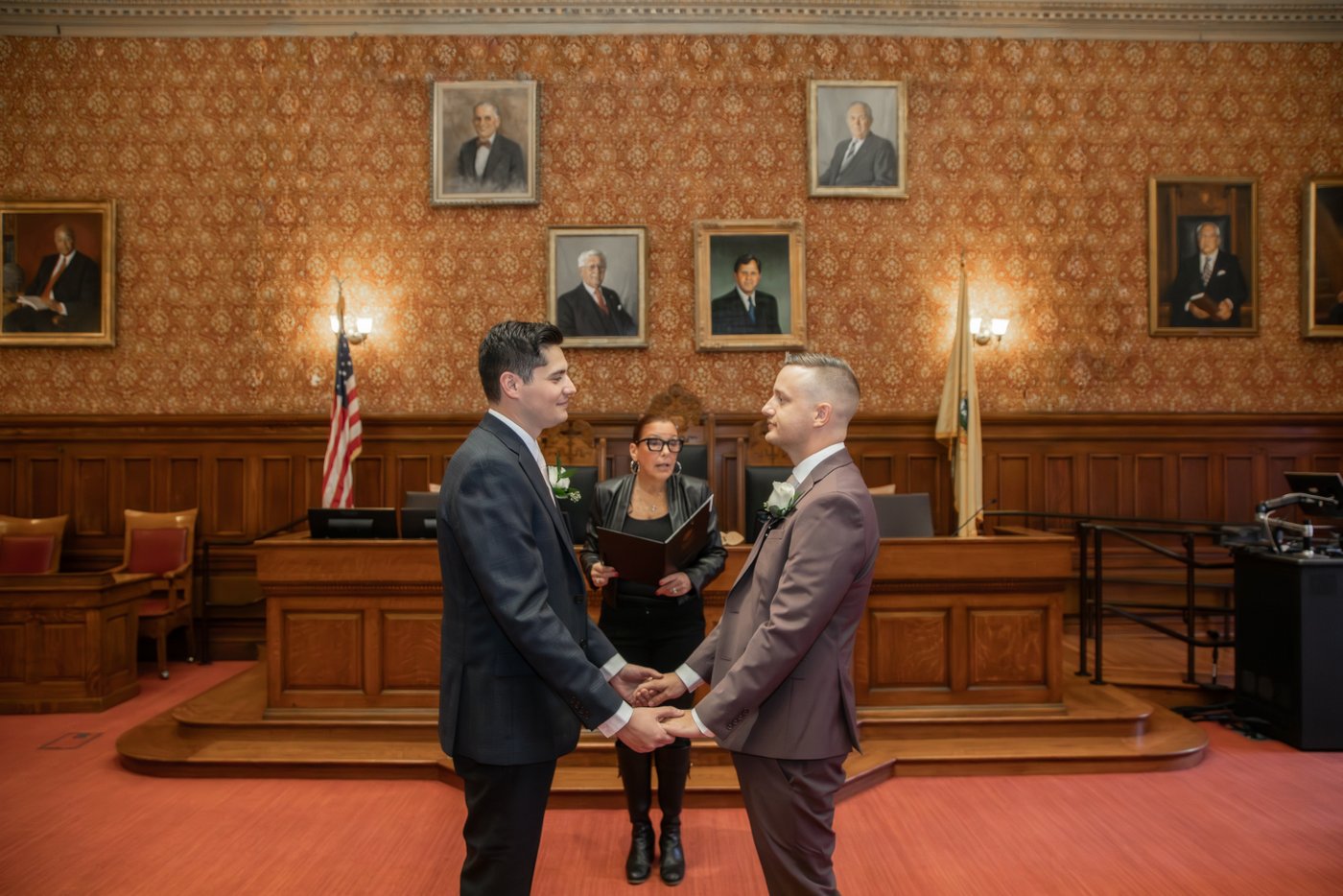 A scene from a Cambridge City Hall wedding ceremony featuring two grooms holding hands as the city clerk officiates their wedding in the Sullivan Chamber.