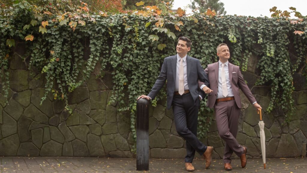 Two grooms dressed in elegant suits stand arm-in-arm in front of an ivy-covered wall in Franklin Street Park, before their Cambridge City Hall elopement
