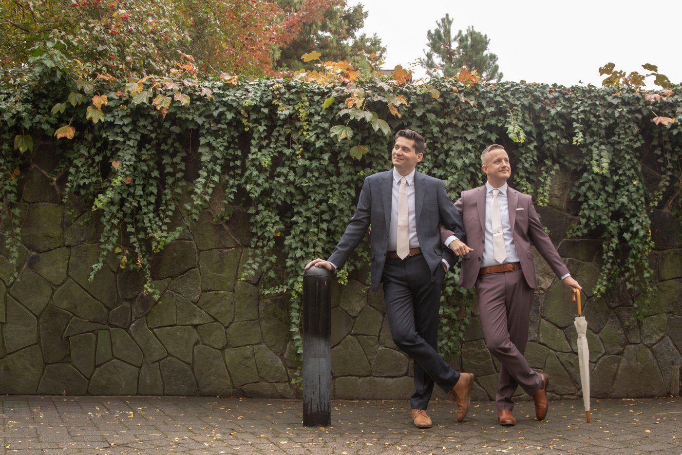 Two men dressed in elegant suits stand arm-in-arm in front of an ivy-covered wall in Franklin Street Park, before their Cambridge City Hall elopement