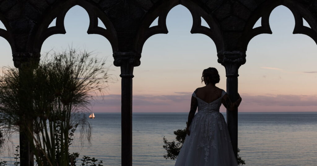 A Hammond Castle Gloucester wedding portrait of a bride looking out at the ocean and a sailboat at sunset