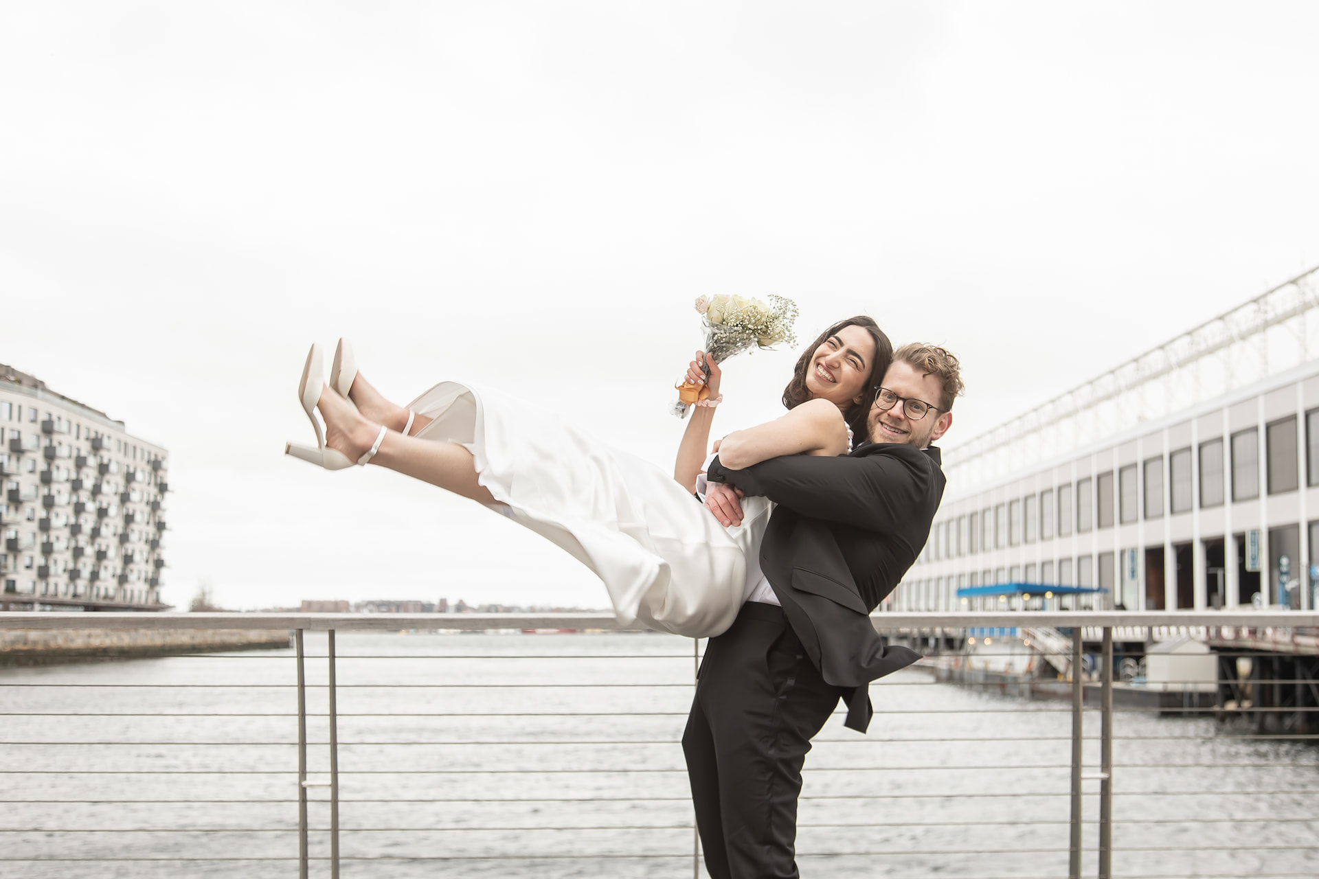 A fun and candid photo of a groom lifting a bride into the air on the Evelyn Moakley Bridge in Boston's Seaport District on their wedding day, featured on Spagnolo Photography's Micro Wedding Photography Packages page.