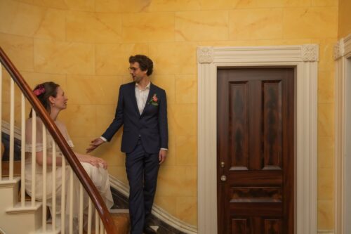 A bride sits on the stairs, the groom leans against a yellow wall next to her, a wooden door next to him in a brownstone building Boston's Back Bay, where they held their micro-wedding - photographedina creative candid style by Spagnolo Photography as part of their Boston micro-wedding photography package.