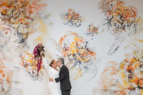 A bride raises her bouquet above her head as she kisses the groom in front of a massive mural of wedding bouquets, following her MIT Chapel micro-wedding, which was shot as part of a micro-wedding package by Spagnolo Photography