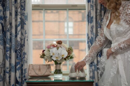 In front of a window framed by blue and white curtains, a small table holds a bouquet, a pair of white shoes, and an off-white satin purse with a pearl handle, as the bride in a lace robe reaches for the shoes - photographed at the bridal suite of the Eliot Hotel by Spagnolo Photography, as part of a micro-wedding coverage