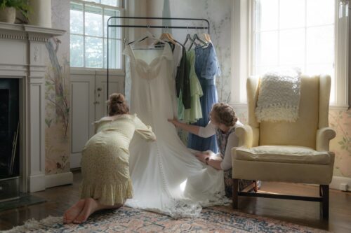 A bride's friends are on their knees fixing the wedding gown that's hanging on a gown in a quaint room with floral wallpaper and natural light at the Long Hill Estate in Beverly, MA, photographed in a candid yet artistic style by creative wedding photographers Spagnolo Photography for a micro-wedding photography package