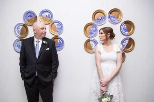 A portrait of a bride and groom standing in front of a white wall a few feet apart, behind them are blue and gold plates hung in a circle that frame their heads like a halo in the lobby of a Boston hotel on their micro-wedding day