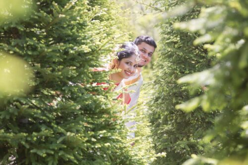 A bride and groom poke their heads out from behind a busk on the grounds of the Sri Lakshmi Temple in Ashland, MA, photographed by Spagnolo Photography as part of their micro-wedding photography package.