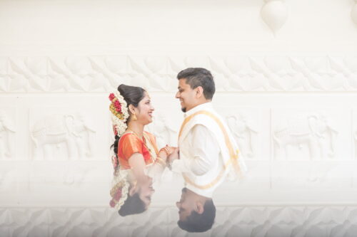 An artistic portrait of a bride in red with flowers in her hair and a groom in white standing in front of a white wall holding hands as they smile at each other, their reflections are mirrored on the marble table in front of them at the Sri Lakshmi Temple in Ashland, MA, photographed by creative wedding photographers Spagnolo Photography for their micro-wedding photography package.