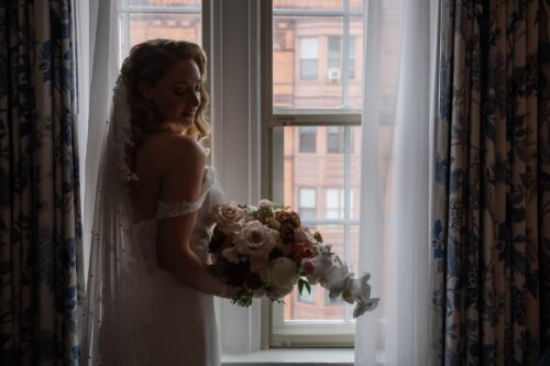 A bride stands by the window of the Eliot Hotel in Boston, a large wedding bouquet in her hands - photographed in natural light by Boston wedding photographers Spagnolo Photography, as part of a micro-wedding photography package.