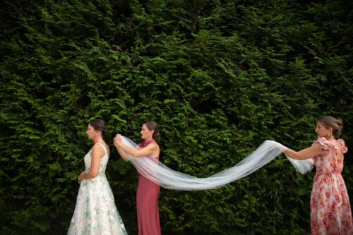 Two bridesmaids in pink dresses roll out the bride's veil and are about to pin it to her hair; the three figures and the veil stand out against the large dark green manicured bush behind them - photographed in Plymouth, MA, by Spagnolo Photography in their creative style as part of the bride's micro-wedding photography package