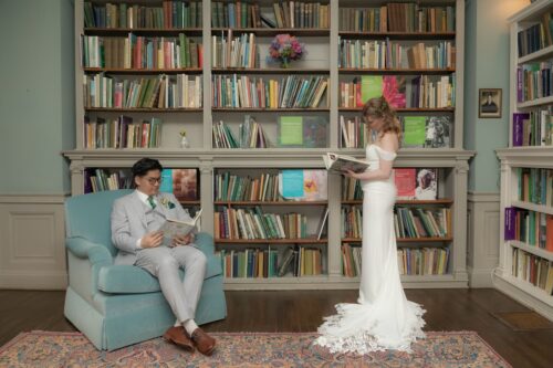 A bride flips through a book while standing and a groom sits in an armchair reading in the library of the Long Hill Estate in Beverly, MA, photographed by Spagnolo Photography for their micro-wedding package