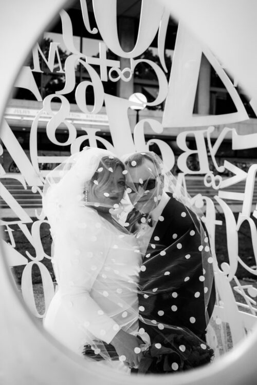 A black and white photo of a bride and a groom wrapped in the bride's polka dotted veil as they stand inside the Alchemist sculpture at MIT in Cambridge, MA - photographed by Spagnolo Photography for their micro-wedding photography package.