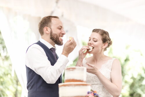 A bright and light photograph of a bride and groom eating their wedding cake while smiling, photographed by Spagnolo Photography for their micro-wedding photography package that included two photographers.