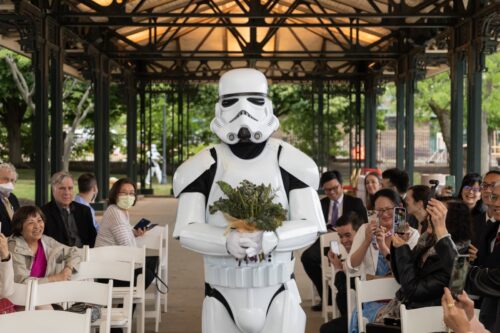 A Star Wars Storm Trooper walks down the aisle holding a bouquet of flowers at an outdoor micro-wedding ceremony in the pavillion of the Museum of Science, photographed by the creative Spagnolo Photography.