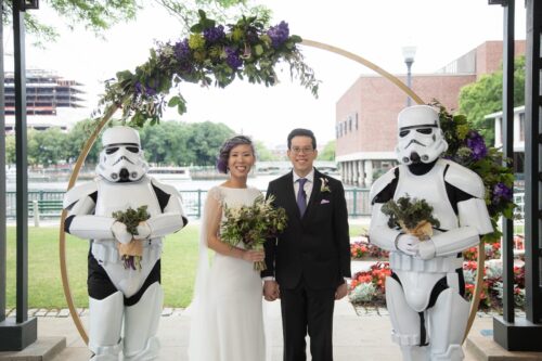 A bride with short purple hair and a white gown and a groom in a black suit stand at the altar decorated with purple flowers and flanked by two Star Wars Storm Troopers acting as their wedding party and holding flower bouquets in the outdoor ceremony space at a Museum of Science wedding, photographed by Spagnolo Photography as part of their micro-wedding package.